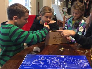 Three children work on brown box on a table using ascrewdriver, next to a large blue schematic