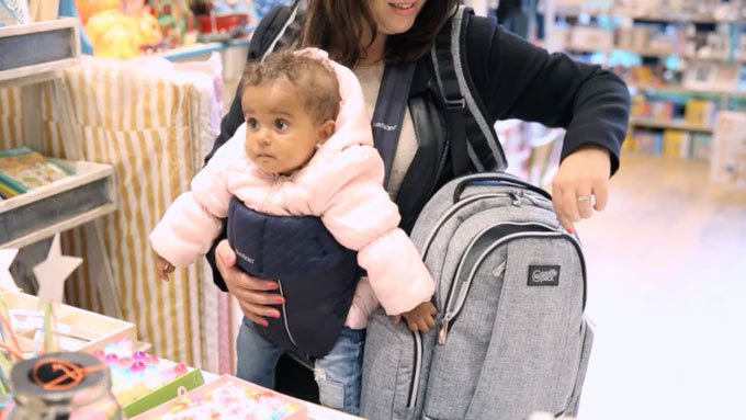 A woman with a baby strapped to her front reaches into a backpack on her side